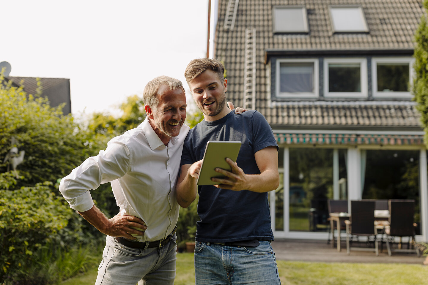 Sohn und Vater stehen im Garten vor einem modernen Einfamilienhaus und betrachten gemeinsam etwas auf dem Tablet.