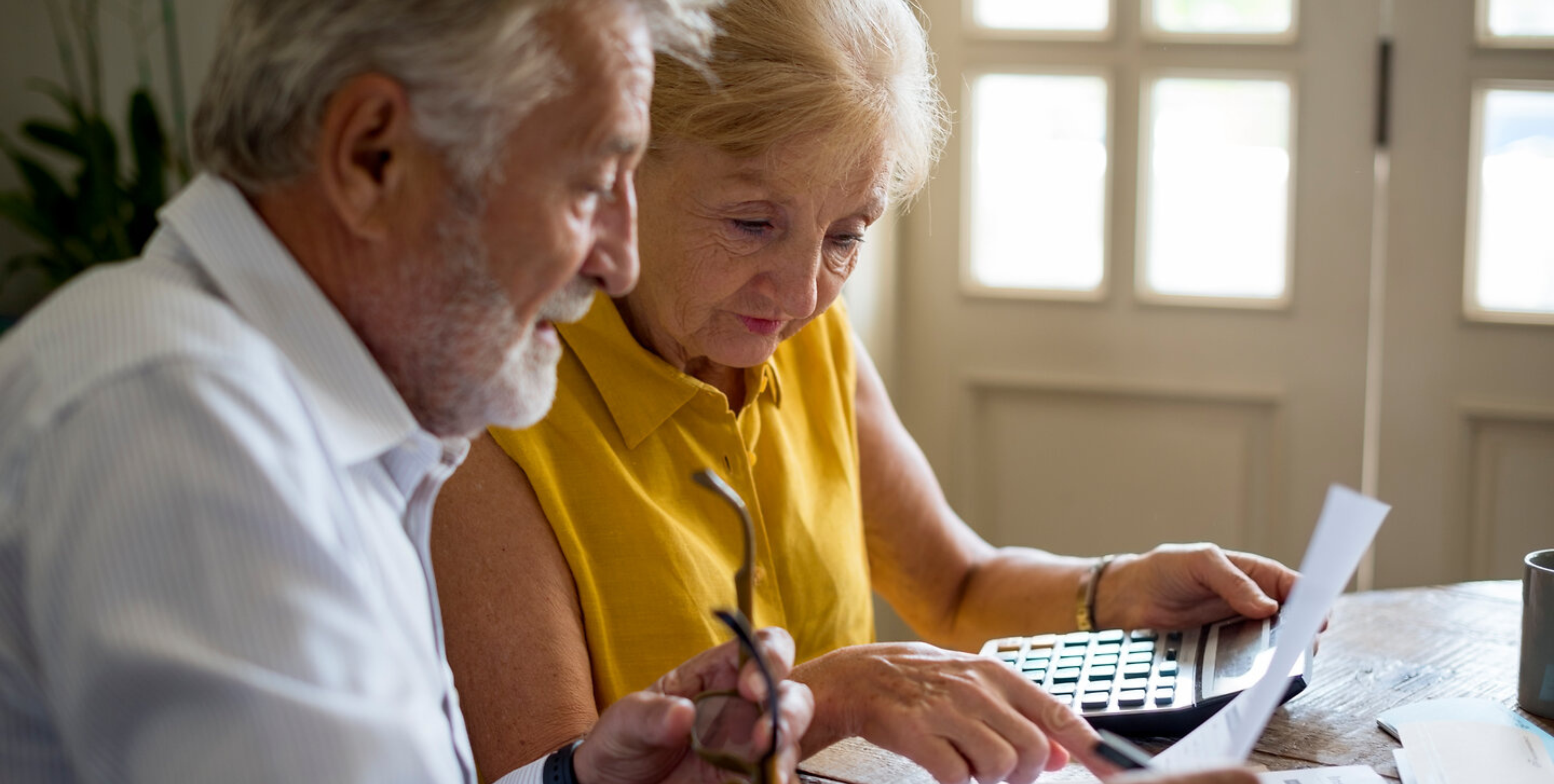 Seitlicher Blick auf älteres Paar, das an einem Tisch sitzt und auf verschiedene Unterlagen schaut. Sie hält einen Taschenrechner in der Hand.