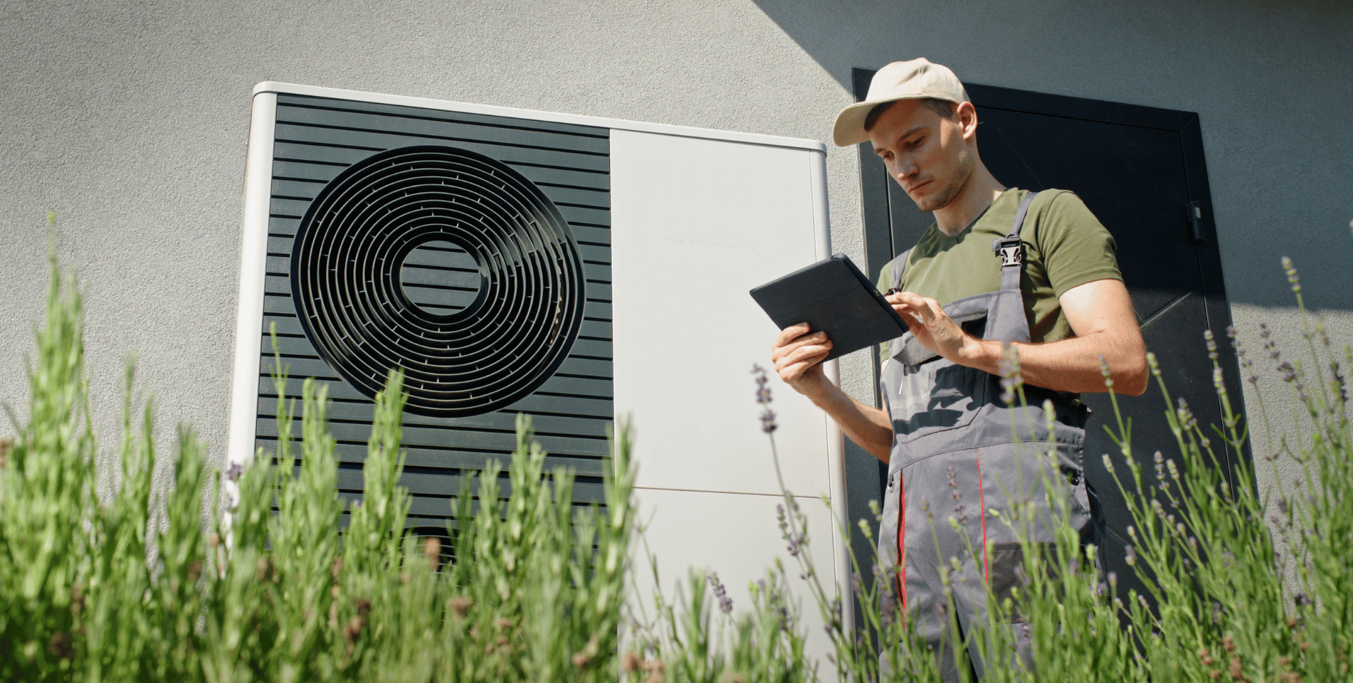 Ein Techniker überprüft mit einem Tablet die Funktion einer Außenwärmepumpe vor einem Wohngebäude bei Sonnenschein.
