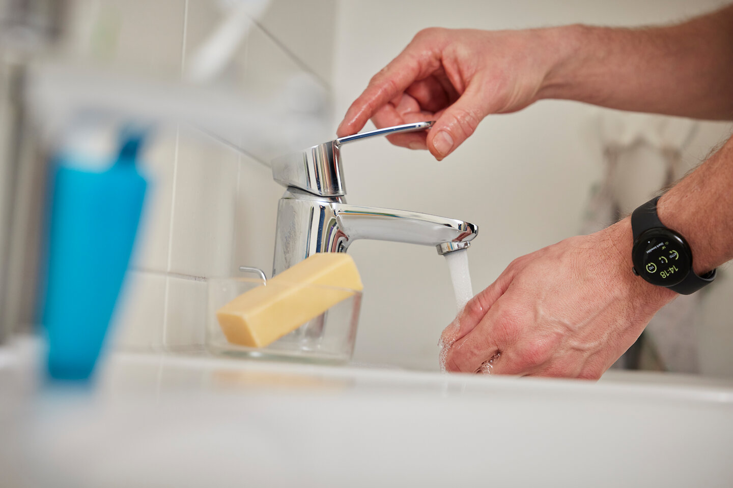 An einem Waschbecken mit Wasserhahn und Seife wäscht sich eine Person die Hände. Man sieht nur die Hände der Person. 