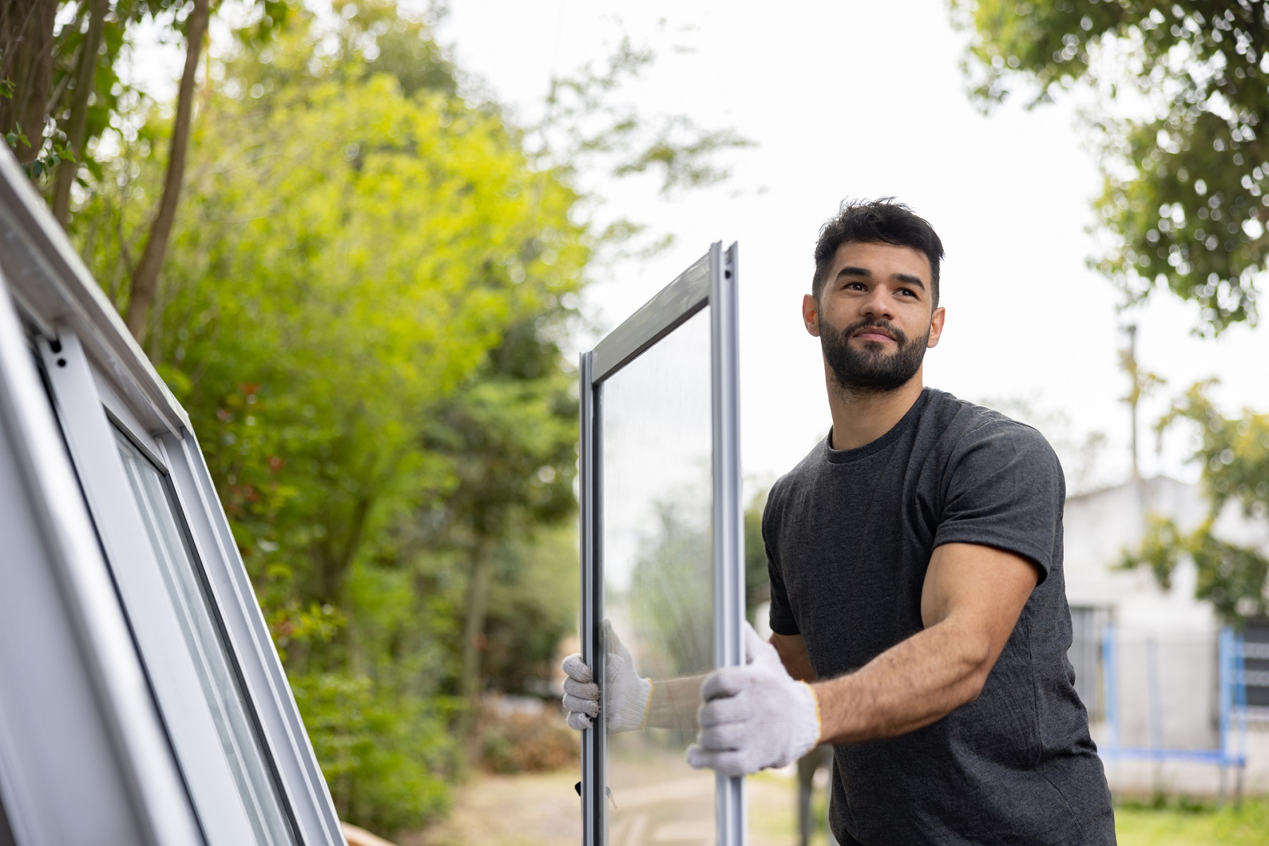 Bauarbeiter mit weißen Schutzhandschuhen trägt einen Teil einer großen Fensterfront oder verglasten Schiebetür vor dem Haus.