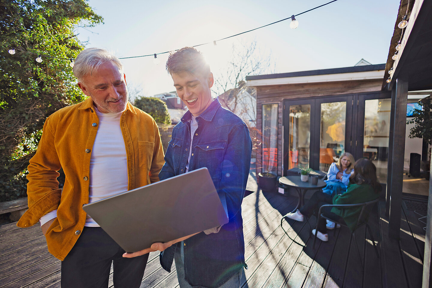 Vater und Sohn schauen im Garten vorm Haus auf einen Laptop, während die Familie in der Nähe ist