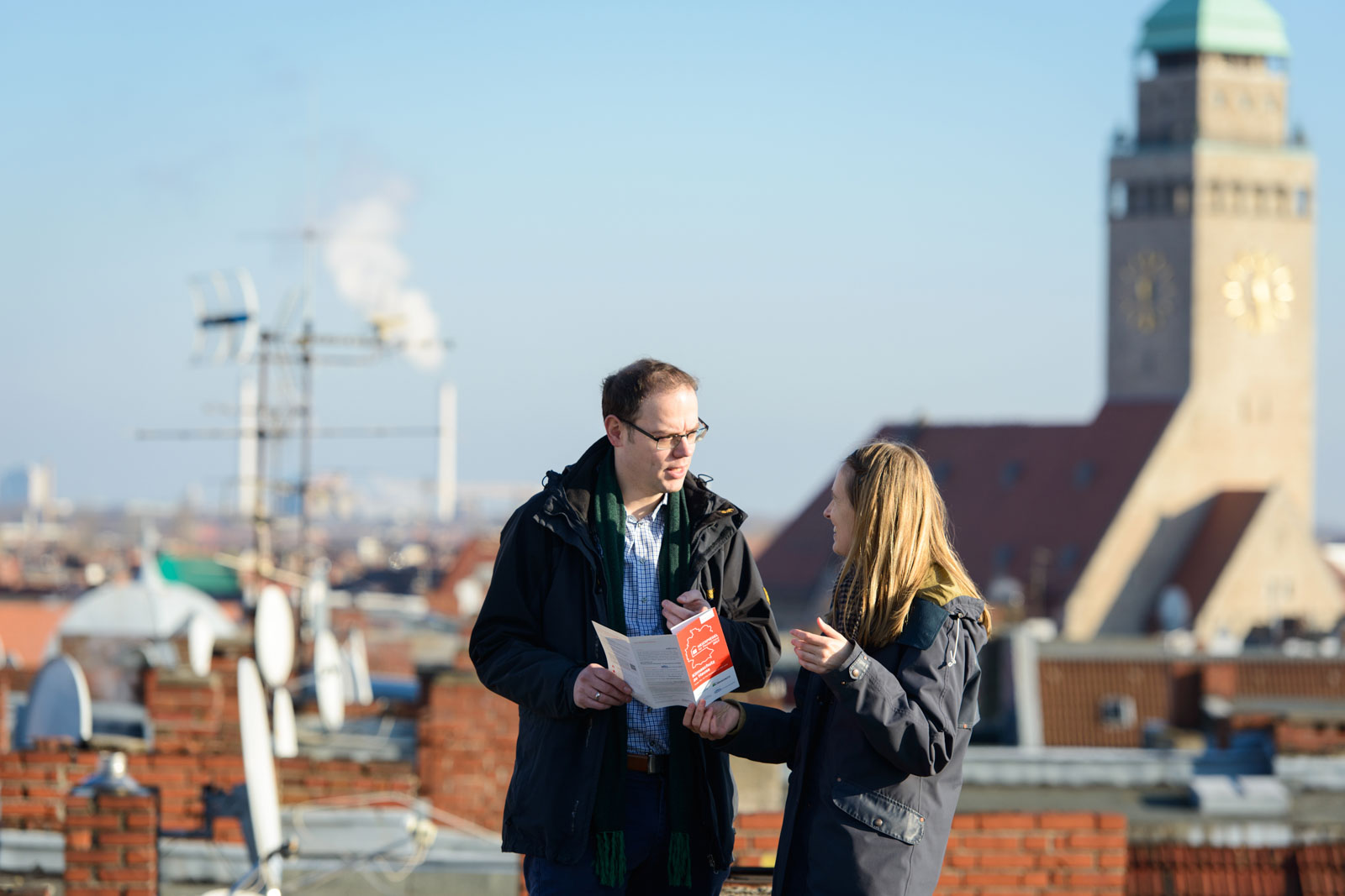 Till Eichmann und eine Frau stehen auf einem Dach und unterhalten sich. Der Mann hat einen Heizspiegel-Flyer in der Hand.