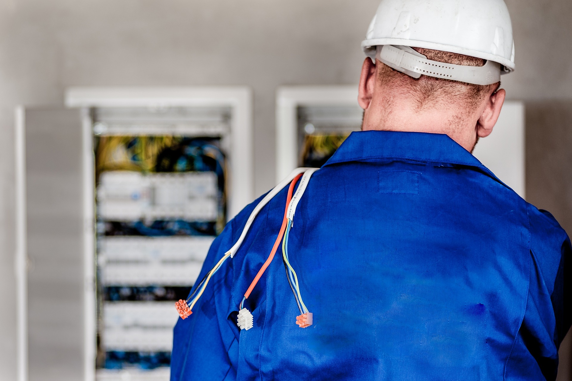Elektriker mit Helm und blauem Anzug vor einem Stromkasten in der Rückenansicht