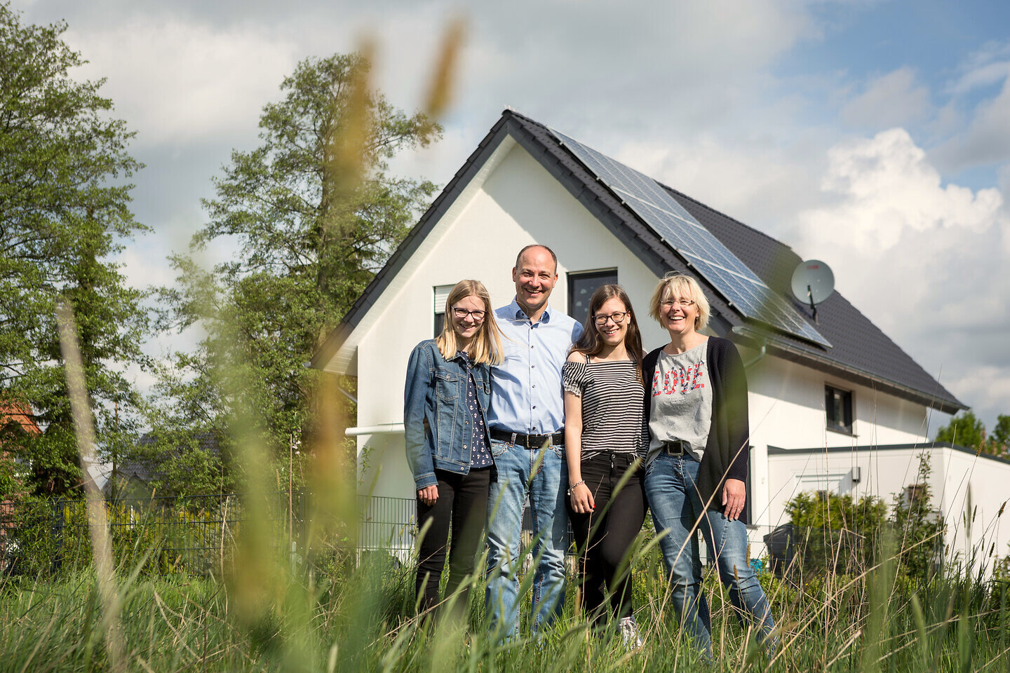 Eine Familie steht im Garten vor ihrem Haus und guckt in die Kamera. Auf dem Dach vom Haus sieht man Photovoltaikanlagen. 