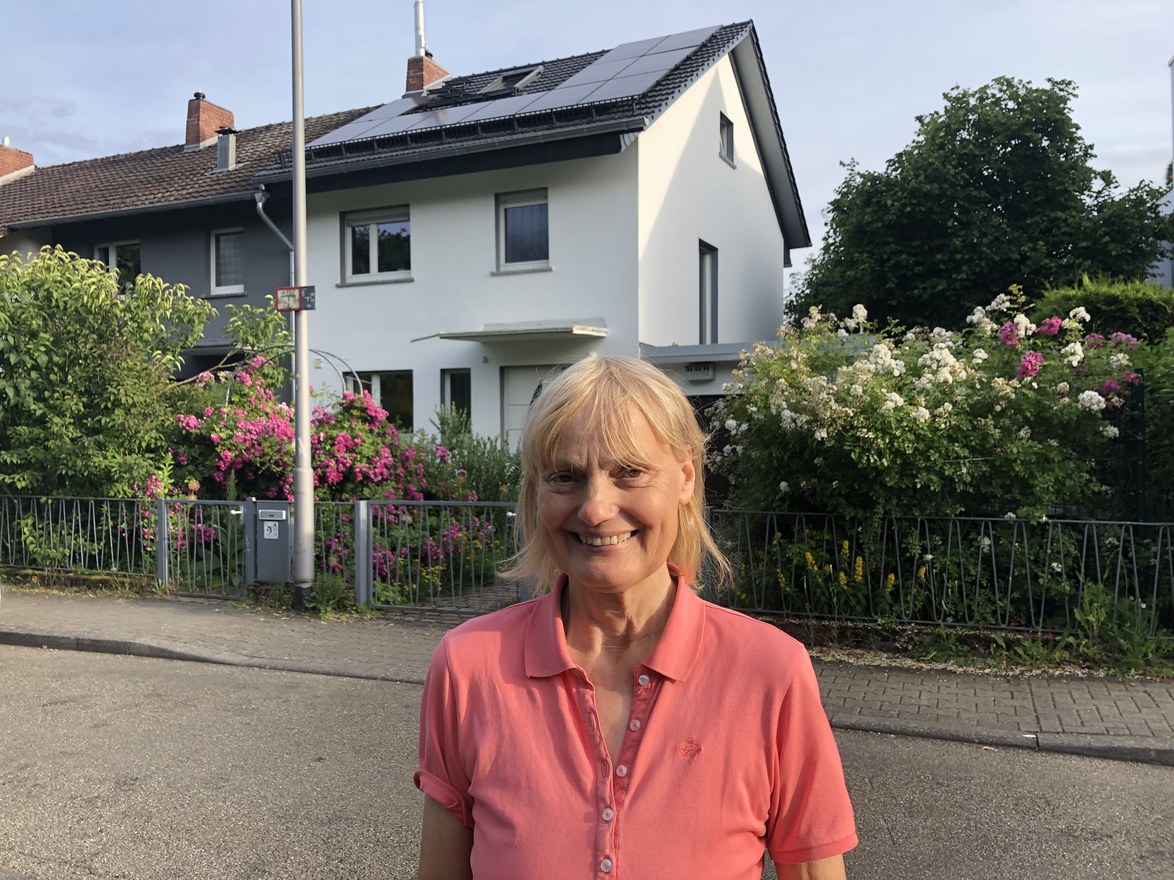 Claudia steht vor ihrem Haus mit Photovoltaikanlagen auf dem Dach und lächelt in die Kamera. 