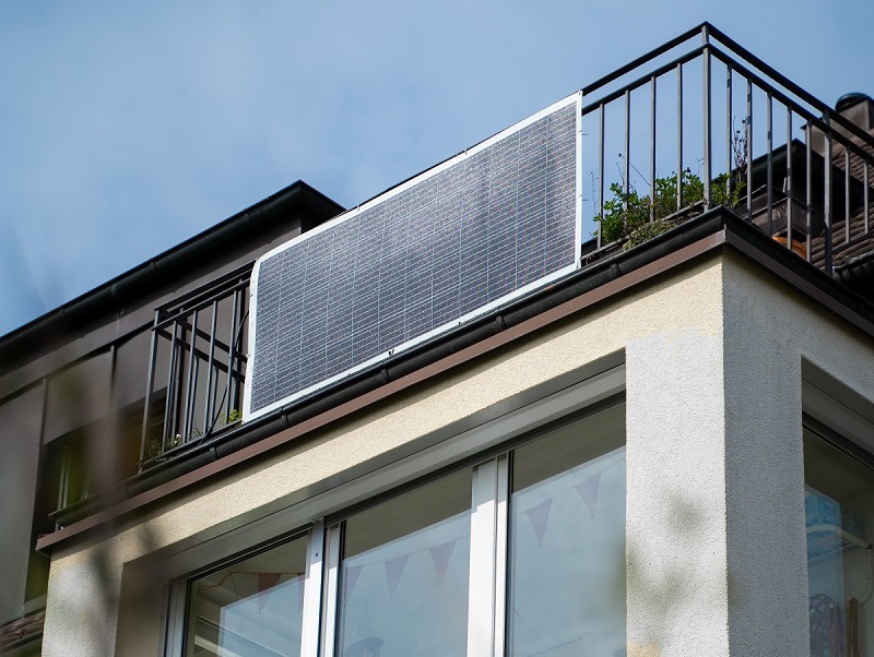 Modernes Mehrfamilienhaus mit einem Balkon im obesten Storckwerk. Auf dem Balkon eine PV-Anlage und eine ältere Dame im blauen Pullover.
