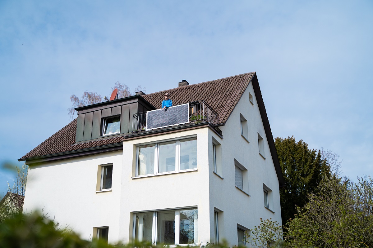 Modernes Mehrfamilienhaus mit einem Balkon im obesten Storckwerk. Auf dem Balkon eine PV-Anlage und eine ältere Dame im blauen Pullover.