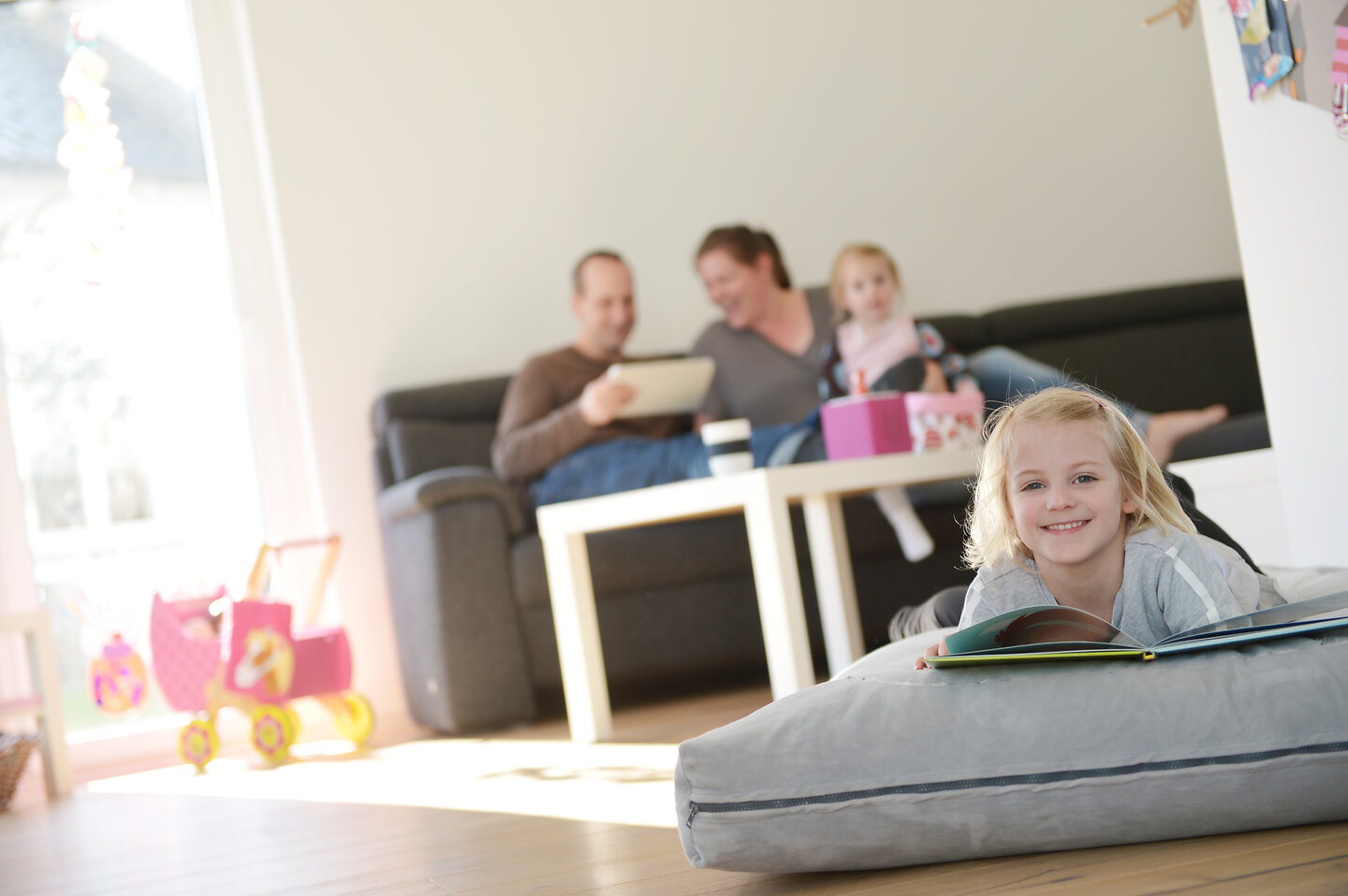 Im Vordergrund liegt ein Mädchen auf einem Kissen, lächelt in die Kamera und hält ein Buch in den Händen. Im Hintergrund sitzen ihre Eltern und ihre Schwester auf der Couch und schauen sich etwas auf einem iPad/ Tablet an