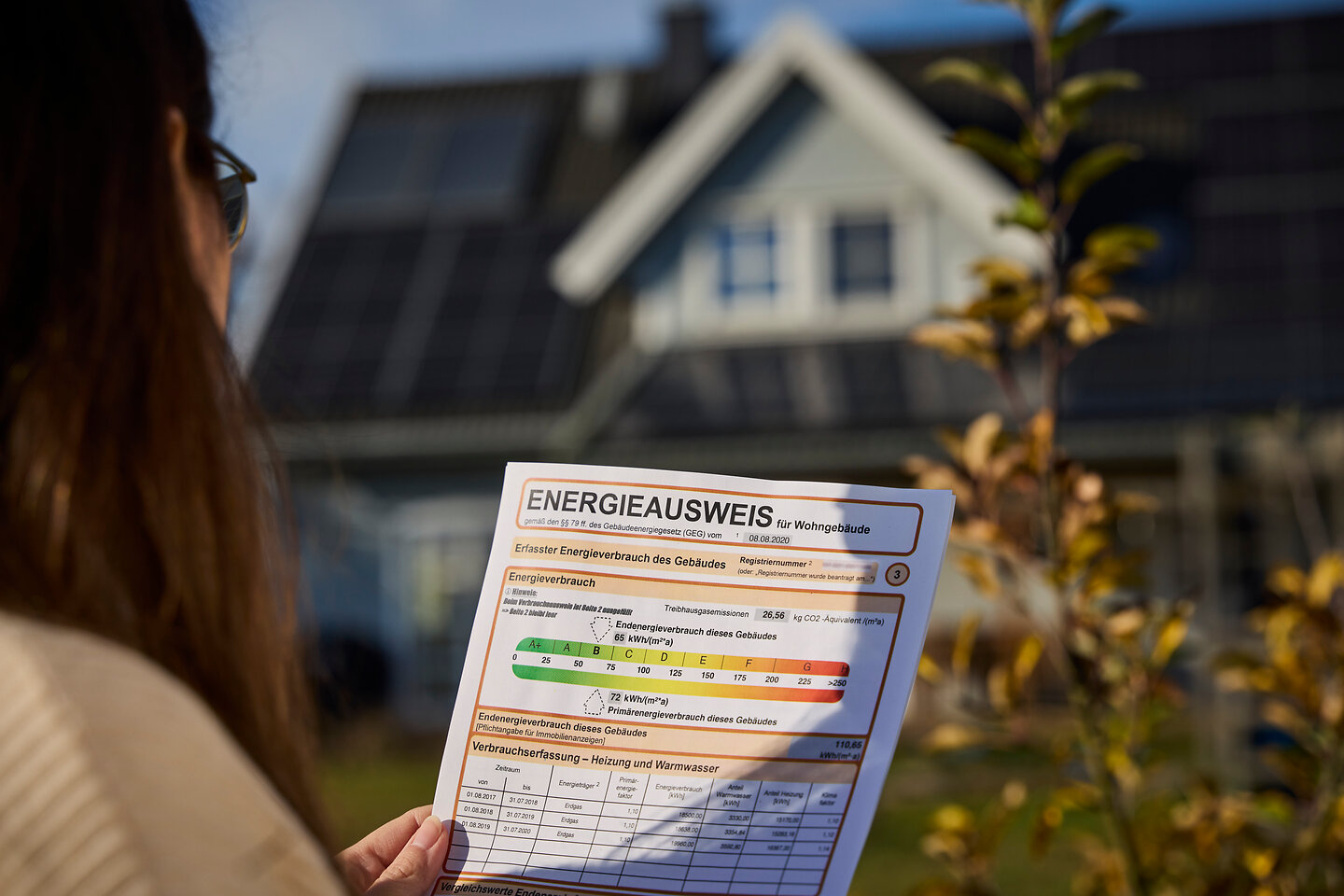 Blick auf die Vorderseite eines Energieausweises, den eine Frau in der Hand hält. Im Hintergrund ein modernes Haus.