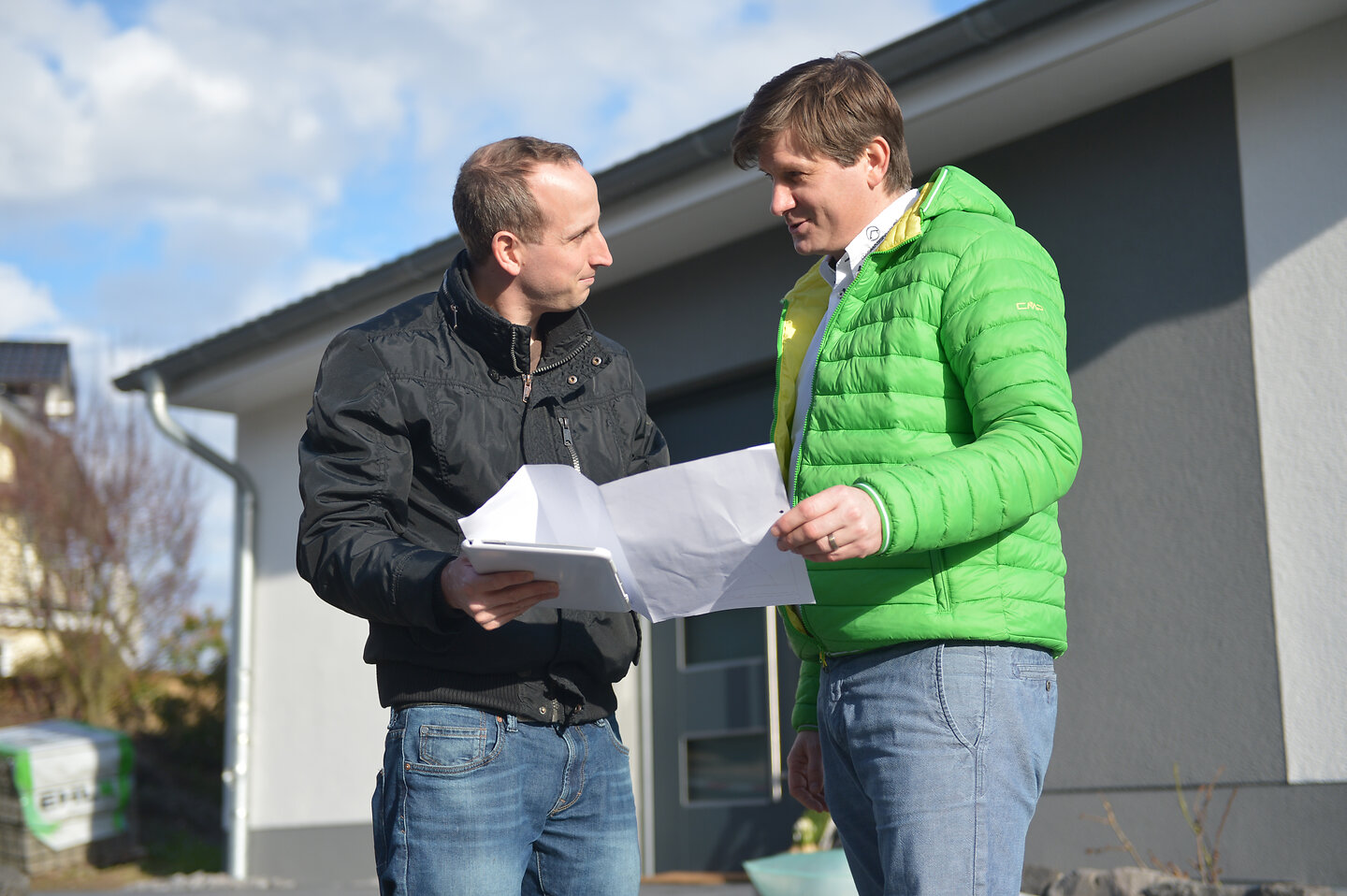 Zwei Männer stehen vor einem Haus und schauen zusammen in Papierunterlagen.