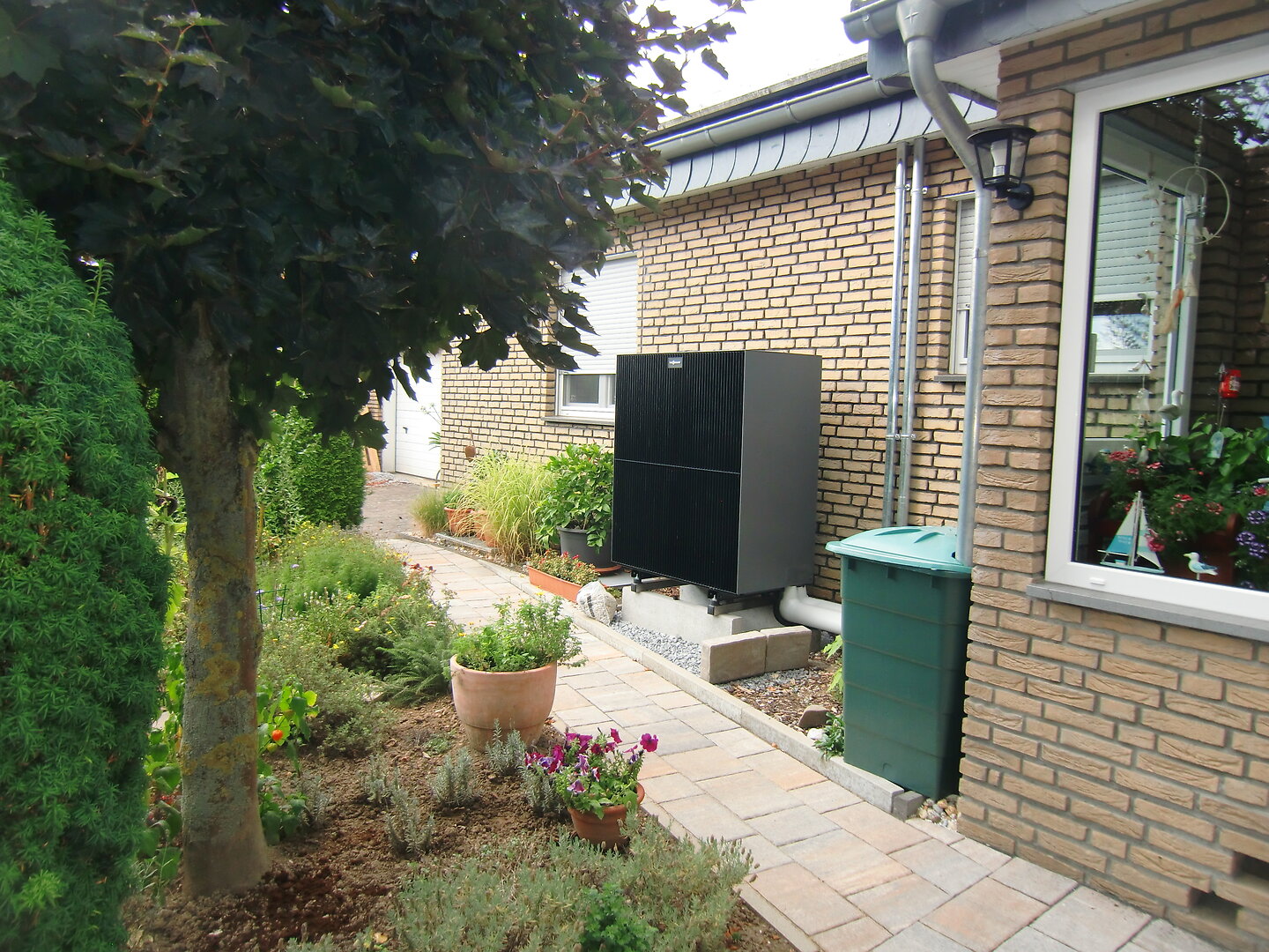 Zu sehen ist ein Haus mit Steinweg und Garten. An der Hauswand steht eine Wärmepumpe. 