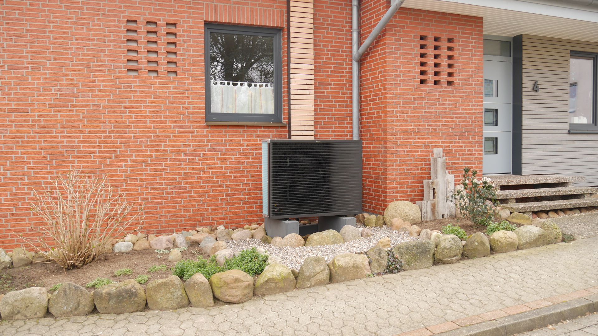 Fassade eines roten Backstein-Hauses mit einer Wärmepumpe im Vorgarten.