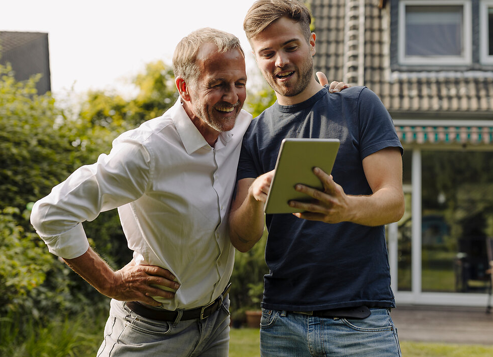 Sohn und Vater stehen im Garten vor einem modernen Einfamilienhaus und betrachten gemeinsam etwas auf dem Tablet.
