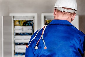 Elektriker mit Helm und blauem Anzug vor einem Stromkasten in der Rückenansicht