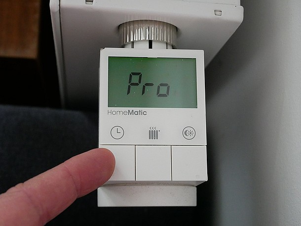 Blick von oben auf Smart-Thermostat von Homematic IP mit grün leuchtendem Display und Anzeige Pro, Zeigefinger auf linker Taste, im Hintergrund weißer Heizkörper und braune und blaue Möbel