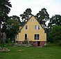 Rasen, Einfamilienhaus mit gelber Fassade und Spitzboden, im Hintergrund Bäume