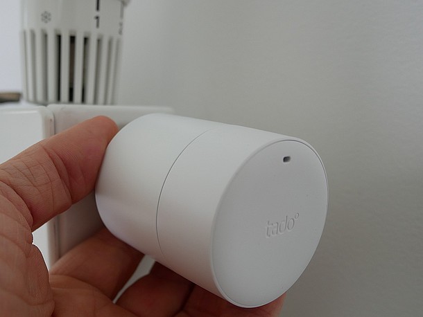 Smartes Thermostat von tado am Heizkörper, links haltende Hand und Finger, im Hintergrund Heizkörper und herkömmliches Thermostat
