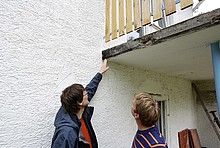 Energieberater und Kunde blicken an einem weißen Gebäude von unten auf einen maroden Balkon mit Holzgeländer