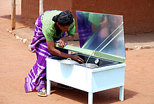 Frau mit Solarkocher vor einer Lehmhütte