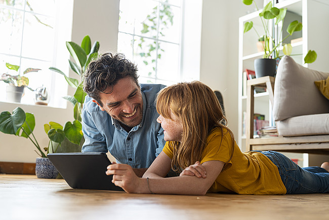 Smiling daughter with digital tablet looking at father while lying on floor in living room at home