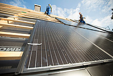 Installation von Photovoltaik auf einem Dach: Module auf Dachlatten von unten fotografiert, oben Schornstein und zwei Handwerker